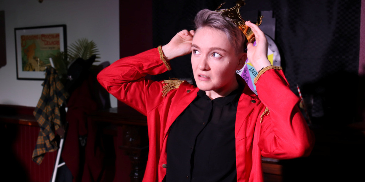 A performer with short, blonde hair in a red blazer looks off to the side in concern while putting a crown on their head.