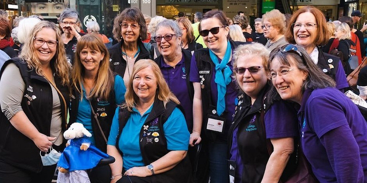 Dare To Dream - A group of 9 women dressed in purple and aqua blue polo shirts smiling at the camera, with shop windows from Rundle Mall in the background