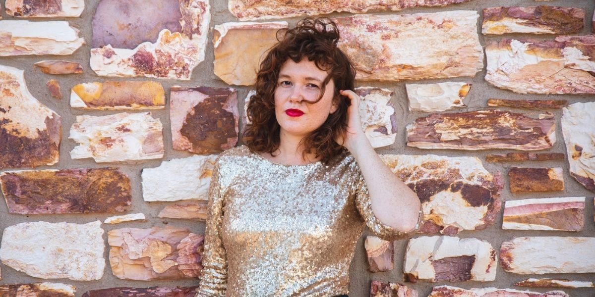 A Caucasian woman in her early thirties stands against a brownstone brick wall. She has short, dark, curly hair blowing in the breeze and is wearing bright red lipstick that pops against her pale skin. She has on a champagne coloured sequin top and is looking directly at the camera.