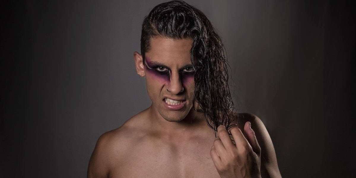 Rouge Goes Rogue - Muscular shirtless man with strong black and pink eye make up snarls looking at the camera, while twisting long, dark, curly hair through his fingers