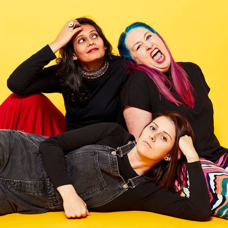 Three women in a seated pose against a yellow background. Kru Harale is South-Asian and wears a black top and red skirt. Leaning against her is Kelly Mac, a Caucasian with pink and blue long hair. Stretched out in front is Annie Boyle, a Caucasian wearing a black skivvy and denim overalls. Kru looks off-camera with a small smile, Kel's mouth is open wide, and Annie stares straight ahead, deadpan.