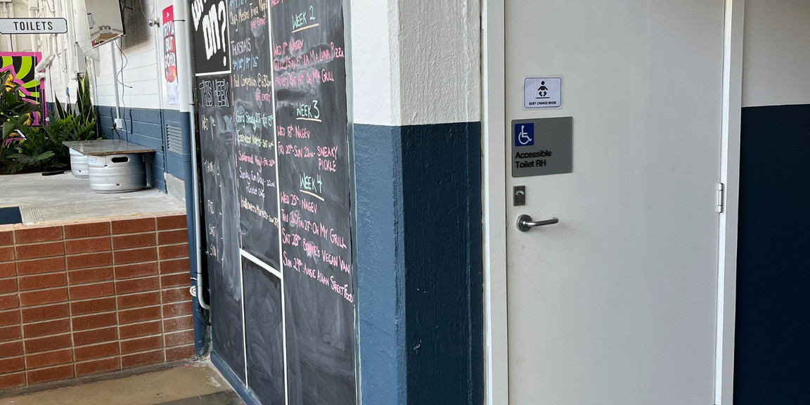 The image is of the accessible bathroom/baby change area door. The bathroom is located in the hallway leading to the office, on the righthand side as you enter the venue. The bathroom is unisex, accessible, and includes a baby-changing station as well as sanitary bins.