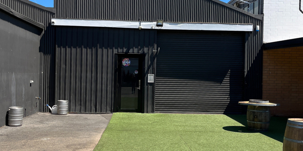 The image is of the main entrance to the venue. There is a glass door in the middle of a warehouse wall and a roller door on the right hand side. The roller door is open when weather is decent meaning there are two points of entry to the space. Leading up to the door the ground is flat and covered in astro turf. There are some outdoor tables on the right as well.